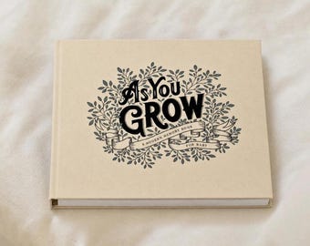 May include: A rectangular memory book with a light tan cover. The front features black text that reads "As You Grow" and "A Modern Memory Book For Baby" surrounded by a detailed illustration of leafy vines and a decorative ribbon banner.