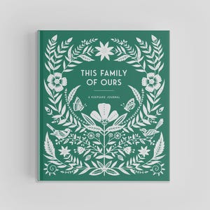 以下が含まれることがあります： 「THIS FAMILY OF OURS」と「A KEEPSAKE JOURNAL」の白文字が入った、緑色のハードカバーのジャーナル。表紙には、蝶や鳥をあしらった白い花と葉のデザインが施されています。