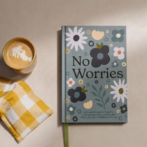May include: A journal with the words "No Worries" on the cover, decorated with floral designs. Beside it are two yellow and white checkered pillows and a cup of coffee with latte art.