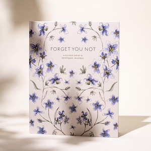 Peut inclure: Un journal &agrave; couverture rigide avec un motif floral de fleurs bleues et violettes et les mots "Forget You Not". Le journal est un journal de deuil guid&eacute; et de souvenirs.