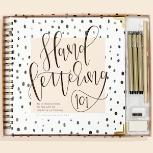May include: A hand lettering kit with a spiral-bound notebook, pens, and accessories. The notebook cover features a polka dot pattern and the words "Hand Lettering 101." Gold-colored pens and a pencil sharpener are included.