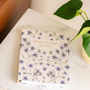 Peut inclure: Un journal &agrave; couverture rigide avec le texte "Forget You Not" et des illustrations florales bleues et blanches. Le journal est un journal de deuil guid&eacute; et de souvenirs. Une plante verte est en arri&egrave;re-plan.