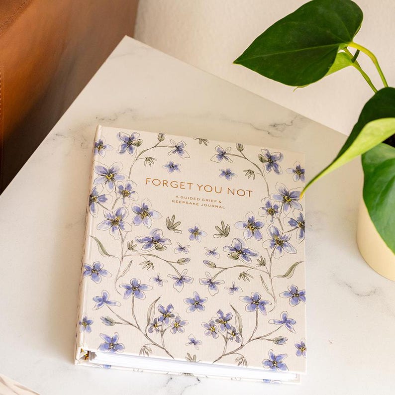 Peut inclure: Un journal &agrave; couverture rigide avec le texte "Forget You Not" et des illustrations florales bleues et blanches. Le journal est un journal de deuil guid&eacute; et de souvenirs. Une plante verte est en arri&egrave;re-plan.