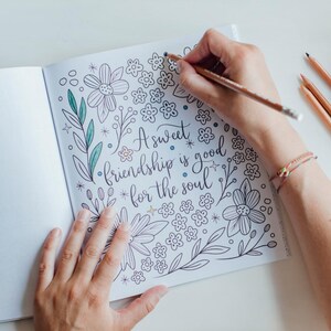 May include: An open coloring book page featuring the hand-lettered text A sweet friendship is good for the soul, surrounded by black and white line art illustrations of various flowers, leaves, and small stars, with some elements partially colored.