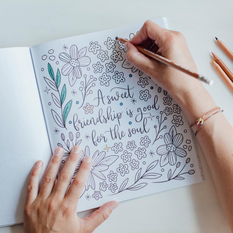 May include: An open coloring book page featuring the hand-lettered text A sweet friendship is good for the soul, surrounded by black and white line art illustrations of various flowers, leaves, and small stars, with some elements partially colored.