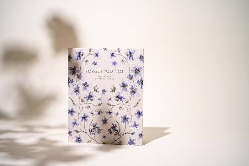 Peut inclure: Un journal &agrave; couverture rigide avec un motif floral de fleurs bleues et violettes et les mots "Forget You Not". Le journal est un journal de deuil guid&eacute; et de souvenirs.