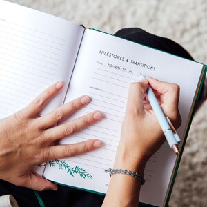 以下が含まれることがあります： 「Milestones & Transitions」の文字と罫線のあるページが開かれた日記。水色のペンで「Homebirth in」と書かれています。日記は緑色の縁取りと花の模様があります。