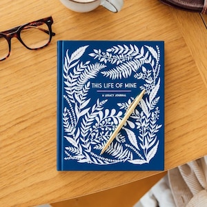 May include: A blue journal with white botanical illustrations and the text "THIS LIFE OF MINE" and "A LEGACY JOURNAL". A gold pen rests on the journal. Eyeglasses and a cup are in the background.