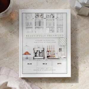 May include: A hardcover book titled Beautifully Organized: A Guide to Function and Style in Your Home by Nikki Boyd. The cover features a photograph of a white kitchen with glass-front cabinets, a coffee maker, glass bottles, and a table lamp.