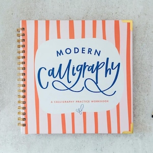 May include: A square spiral-bound workbook with a light pink cover featuring vertical orange stripes. The center displays the text "MODERN CALLIGRAPHY" in blue, followed by "A CALLIGRAPHY PRACTICE WORKBOOK" and a small blue monogram logo.