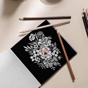 May include: A black coloring book page with a white floral design. The page is open and lying on a white surface. There are several colored pencils on the surface, including brown, beige, and peach.