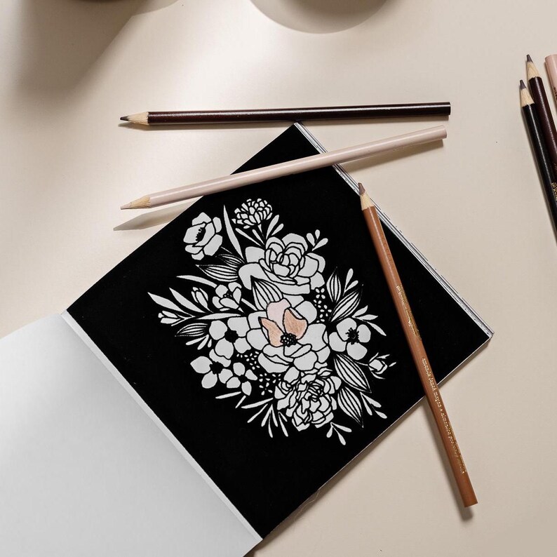 May include: A black coloring book page with a white floral design. The page is open and lying on a white surface. There are several colored pencils on the surface, including brown, beige, and peach.