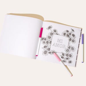 May include: An open coloring book with a floral design and the words "Mi Amor" in the center. Three colorful markers, pink and purple, are placed near the book.