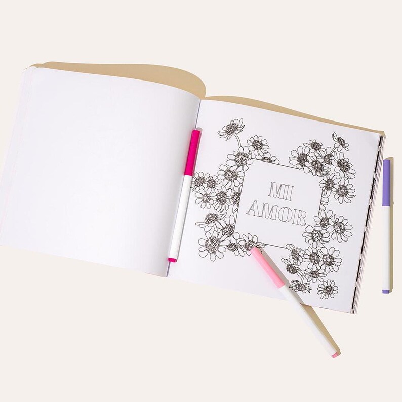 May include: An open coloring book with a floral design and the words "Mi Amor" in the center. Three colorful markers, pink and purple, are placed near the book.