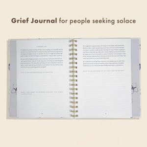 Peut inclure: Journal de deuil ouvert avec une reliure &agrave; spirale dor&eacute;e. Les pages du journal sont remplies de texte et d'invites. La couverture et les pages ont un motif floral dans les tons de bleu et de violet. Le texte en haut indique "Grief Journal for people seeking solace."