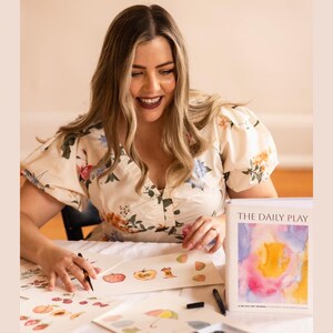 A picture of author, Brittany DeSantis holding a paper with water color paintings on it. Her book, The Daily Play is sitting next to her on the table.