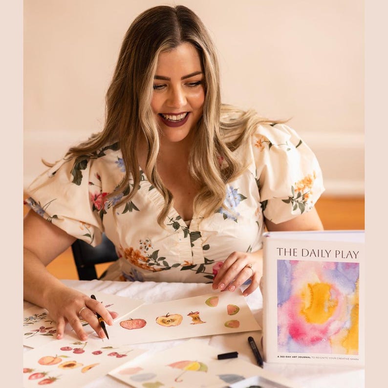 A picture of author, Brittany DeSantis holding a paper with water color paintings on it. Her book, The Daily Play is sitting next to her on the table.