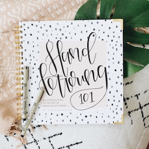 May include: A white and black polka dot notebook with a gold spiral binding. The cover features the text "Hand Lettering 101" in black cursive lettering. The text "An introduction to the art of creative lettering" and "By Chalkfulloflove" is printed in black below the title.