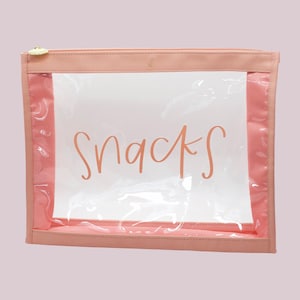May include: A clear, rectangular pouch with a pink border and a gold zipper. The word "SNACKS" is written in a cursive font on a white insert. The pouch is designed for storing snacks and other small items.