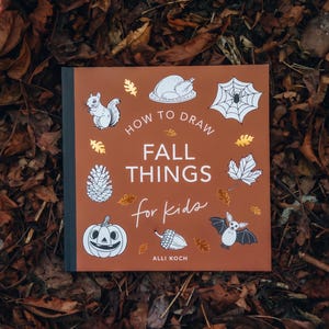 May include: A square book with a rust-colored cover titled How to Draw Fall Things for Kids by Alli Koch. The cover features white line illustrations of a squirrel, turkey, spider web, pinecone, maple leaf, jack-o'-lantern, acorn, and bat.