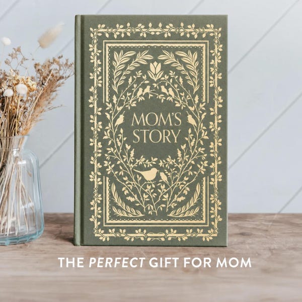 May include: A hardcover book with the title "MOM'S STORY" in gold lettering. The cover is olive green with a gold floral and bird design. The book is standing upright on a wooden surface.