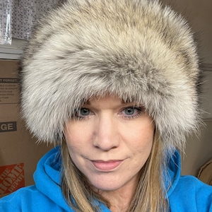 May include: A woman wearing a fluffy white fur hat. The hat is large and covers most of her head. She is wearing a blue hoodie.