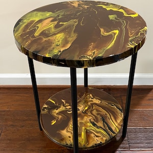 May include: A round side table with a two-tiered design. The tabletop and lower shelf feature a brown, yellow, and black marbled pattern. The table has black metal legs and measures approximately 20 inches in diameter and 20 inches tall.