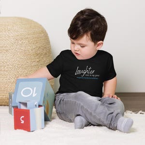 May include: A toddler wearing a black t-shirt with the text "Laughter runs in my genes Angelman Syndrome Awareness" in white. The toddler is sitting on a light brown rug and playing with blue and red blocks.
