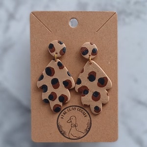 May include: A pair of dangle earrings with a leopard print design. The earrings are made of clay and have gold hooks. The earrings are on a brown card with the text "The Clay Goose" in a circle.