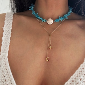 Könnte beinhalten: Ein geschichtetes Halsketten-Set bestehend aus einem Choker aus kleinen, leuchtend türkisfarbenen Steinfragmenten mit einer zentralen weißen Spiralmuschelperle sowie einer feinen goldfarbenen Kette mit einem vierzackigen Stern und einem Mondsichel-Anhänger.