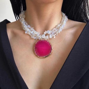 Könnte beinhalten: Eine auffällige Halskette mit einem großen, leuchtend pinkfarbenen Achat-Anhänger mit goldfarbenem Rand. Der Anhänger hängt an einer mehrsträngigen Halskette aus klaren und weißen Perlen. Der Hintergrund ist dunkel.