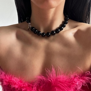 May include: A black beaded choker necklace with a silver clasp. The necklace is worn by a person with long black hair and a pink fuzzy top.