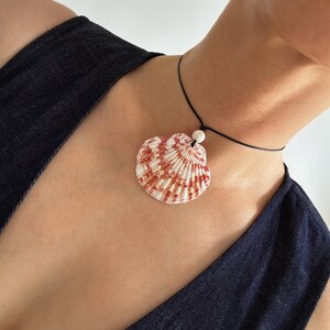 May include: A necklace featuring a single fan-shaped seashell pendant with white and reddish-brown radial stripes. The shell is suspended from a thin black cord and accented with a small, round, textured white bead positioned above the shell.