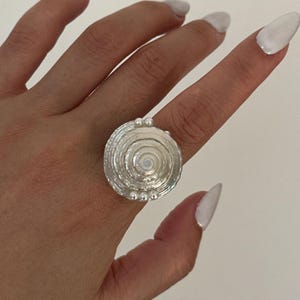 Puede incluir: Un anillo plateado con un diseño en espiral. El anillo presenta una cara circular texturizada con una serie de crestas concéntricas. Pequeñas cuentas redondas están dispuestas alrededor del borde del anillo. El anillo se lleva en un dedo.