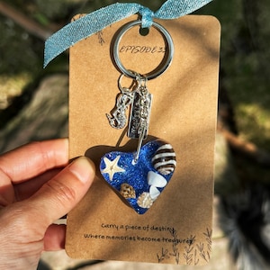 May include: A blue heart-shaped resin keyring with seashells and a starfish inside. The keyring is attached to a silver key ring with a mermaid charm and a charm that says "Strength". The card reads "Carry a piece of destiny where memories become treasure".