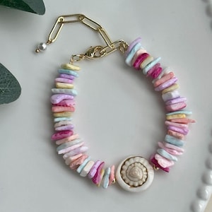 May include: A colorful bracelet featuring a gold chain and clasp. The bracelet is adorned with pastel-colored, irregular-shaped beads in shades of pink, blue, yellow, and purple. A white seashell-shaped charm is centered on the bracelet.