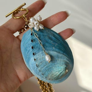 May include: A necklace featuring a large, oval-shaped blue abalone shell pendant attached to a gold-toned toggle clasp. The shell is accented with a cluster of small white pearls and a delicate gold chain drop ending in a single pearl bead.