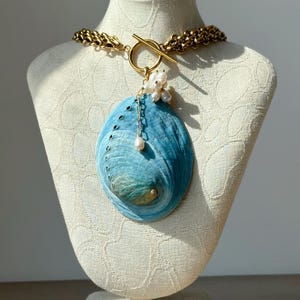 May include: A necklace featuring a large, oval-shaped blue abalone shell pendant suspended from a thick gold-toned chain. The pendant is accented with a cluster of small white freshwater pearls and a single pearl drop attached to a gold toggle clasp.