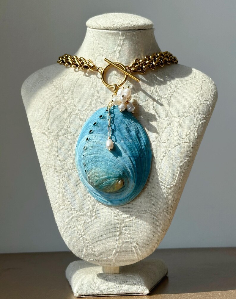 May include: A necklace featuring a large, oval-shaped blue abalone shell pendant suspended from a thick gold-toned chain. The pendant is accented with a cluster of small white freshwater pearls and a single pearl drop attached to a gold toggle clasp.