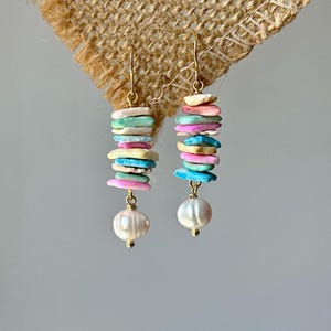 Op de afbeelding: Hangende oorbellen met goudkleurige haken. Elke oorbel heeft een stapel pastelkleurige, platte, ovale kralen in roze, blauw, geel en groen. Aan de onderkant van elke oorbel hangt een grote, witte parel.