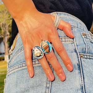 May include: Two silver rings with blue and white seashells. The rings are on a person's finger.