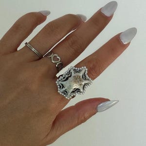 May include: A hand showing three rings: a simple silver band, a ring with heart-shaped cutouts, and a large star-shaped ring. The star ring has a white and black pattern with a pearl edge. The nails are painted a light grey colour.