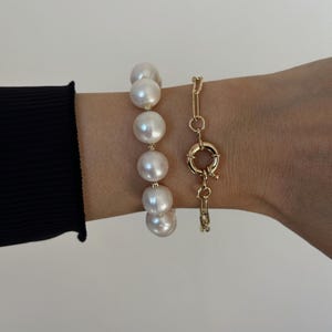 May include: Two bracelets are shown. One features large, round, white pearls. The other is a gold-coloured chain bracelet with a large circular clasp. The bracelets are worn on a wrist, with a black sleeve visible.