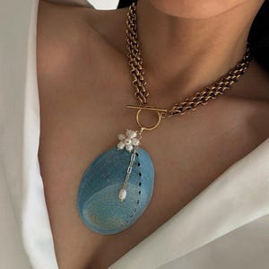 May include: A gold-toned chain necklace featuring a toggle clasp and a large, oval-shaped blue abalone shell pendant. The pendant is accented with a small cluster of white freshwater pearls arranged in a flower shape and a single dangling pearl.
