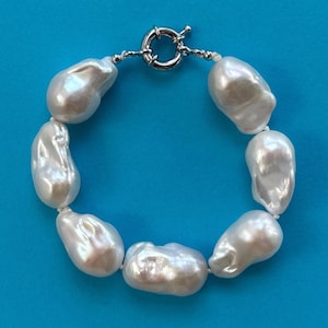 May include: A white pearl bracelet with a silver clasp. The bracelet features large, irregularly shaped pearls strung together. The clasp is a silver toggle style. The pearls have a lustrous sheen, reflecting light.