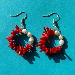 May include: A pair of dangle earrings featuring circular hoops composed of vibrant red coral branch segments and smooth white freshwater pearls. The earrings are suspended from gold-toned metal hooks, creating a contrasting color palette.