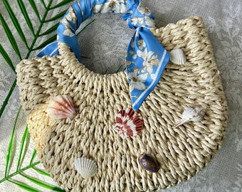 May include: A woven straw handbag with a semi-circular shape, featuring a handle wrapped in a light blue silk scarf with a white floral pattern. The exterior of the bag is decorated with five assorted seashells of varying shapes and colors.