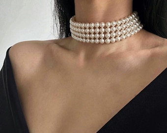 May include: A white pearl choker necklace with multiple strands of pearls. The necklace is worn by a person with dark hair and a black top.