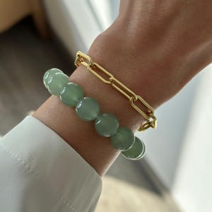 Puede incluir: Un brazalete de cadena dorada y un brazalete de cuentas verdes apilados en la muñeca de una persona.