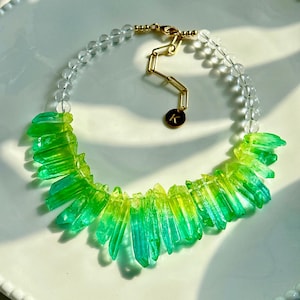 Könnte beinhalten: Eine grüne und blaue Kristallkette mit einer goldenen Kette und klaren Perlen. Die Kristalle haben die Form von langen, dünnen Spitzen.